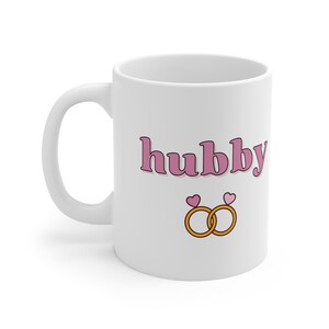 Puede incluir: Taza de cerámica blanca con la palabra "hubby" en letras rosas y dos anillos dorados con corazones rosas en la parte superior.