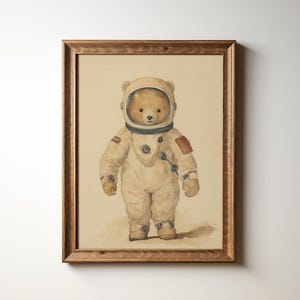 May include: A framed illustration of a teddy bear wearing a white astronaut suit with a brown helmet. The bear is standing on a white background.