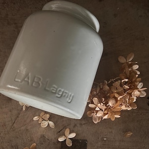 Puede incluir: Un tarro de cerámica blanco con el texto "LAB Lagny" en relieve en el lateral. El tarro está ligeramente inclinado y descansa sobre una superficie de madera, acompañado de flores secas. La estética general es rústica y vintage.