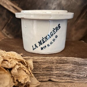 Puede incluir: Un tarro cilíndrico de cerámica blanca con tapa a juego. El lateral presenta un texto impreso en azul oscuro que dice LA MÉNAGÈRE y Bté S.G.D.G. El recipiente tiene un borde plano con pequeñas asas que sobresalen en los lados.