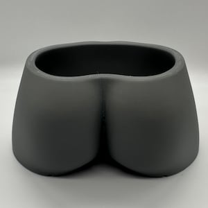 May include: A grey, 3D-printed planter in the shape of a pair of buttocks. The planter has a smooth, matte finish and a wide opening for plants. This unique design is a humorous addition to any home decor.