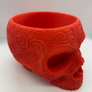 Puede incluir: Un macetero rojo brillante en forma de calavera con un patrón de espiral. El macetero está hecho de un material liso, similar al plástico.