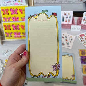May include: A lined notepad with a light blue background and a yellow border. The notepad features a whimsical design with a ladybird, caterpillars, and a butterfly. The word "notes..." is printed at the top. Other stationery items are visible in the background.