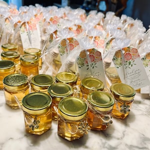 May include: Small glass jars filled with honey, tied with twine and a small gold charm. The jars are arranged on a white surface with a tag that says "Thank you for celebrating!"