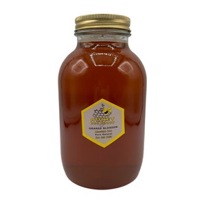 May include: A glass jar of orange blossom honey with a gold lid. The label reads "Honey Bee Happy" and "Orange Blossom" with a cartoon bee illustration. The jar is 500 ml.