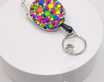Puede incluir: Un portatarjetas de identificación colorido con una vibrante variedad de pedrería en tonos rosa, naranja, verde, azul y morado. El carrete tiene un mosquetón plateado y una correa de plástico transparente con un broche metálico. El portatarjetas está sobre una superficie blanca.