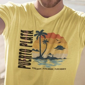 以下が含まれることがあります： ヤシの木と夕焼けのレトロなグラフィックがプリントされた黄色のTシャツ。シャツの左側に「PUERTO PLATA」という文字が縦にプリントされています。「PLAYA SOSUA | PLAYA DORADA | PLAYA CABARETE」という文字がグラフィックの下にプリントされています。