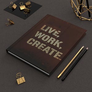 Könnte beinhalten: Ein braunes, ledergebundenes Notizbuch mit den Worten "LIVE, WORK, CREATE" in weiß auf dem Einband. Das Notizbuch liegt auf einer dunkelgrauen Oberfläche mit goldenen Büroklammern und Bleistiften.