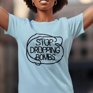 May include: Light blue t-shirt with the text "STOP DROPPING BOMBS" in a black, bubble-like design. The shirt has short sleeves and a crew neck. The text is centered on the front of the shirt.