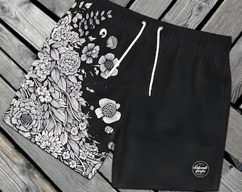 May include: Black swim trunks with a white floral pattern on the left side. The right side is solid black with a white circle logo that says "Seaworthy Surfer".