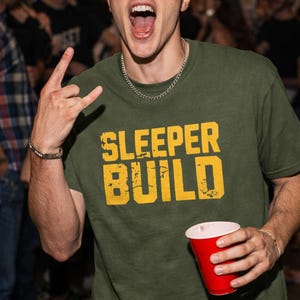 Puede incluir: Camiseta verde oliva con el texto "SLEEPER BUILD" en amarillo, un collar de cadena plateada y un vaso de plástico rojo. La persona está haciendo un gesto con la mano.