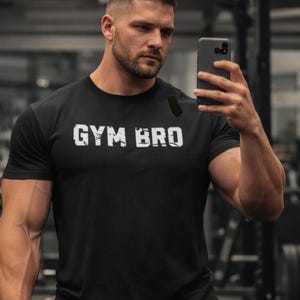 Puede incluir: Camiseta negra de cuello redondo con el texto "GYM BRO" en letras blancas. La camiseta es de manga corta y parece estar hecha de un material suave. La persona de la imagen sostiene un smartphone.