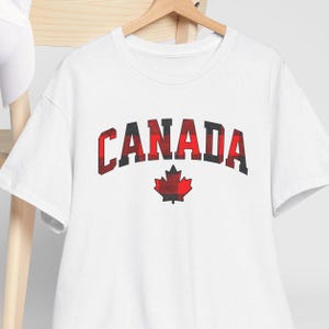 May include: White t-shirt with a red and black plaid design of the word "CANADA" and a maple leaf.