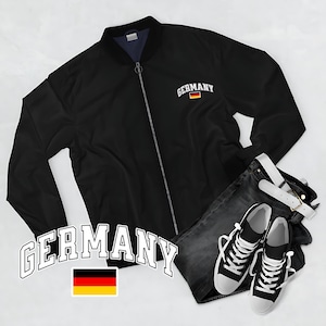 May include: Black bomber jacket with "Germany" text and a German flag patch. Paired with dark wash jeans, a white belt, and black and white sneakers. The jacket has a zipper closure.