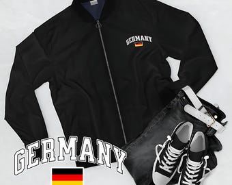 May include: Black bomber jacket with "Germany" text and a German flag patch. Accompanied by dark wash jeans, a white belt, and black and white trainers. The jacket has a zip closure.