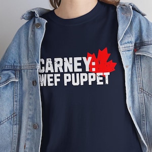 May include: Navy blue t-shirt with the text "CARNEY: WEF PUPPET" in white, and a red maple leaf graphic. The shirt is worn with a denim jacket.