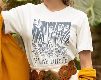 May include: White t-shirt with a blue floral print and the text "PLAY DIRTY" on the front. The shirt is being worn by a person with a brown belt.