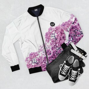May include: White bomber jacket with a black zipper and a floral print of pink flowers. The jacket has a black collar and cuffs. The jacket is laid out on a white surface with a pair of black jeans and black sneakers.