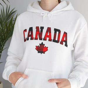 Peut inclure: Sweat-shirt à capuche blanc avec un érable canadien en tissu écossais rouge et noir et le mot "CANADA" en lettres noires et rouges.