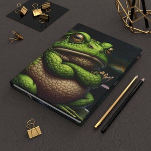 Peut inclure: Un carnet noir avec une illustration de grenouille verte sur la couverture. La grenouille est assise les bras croisés et a une expression sérieuse. Le carnet est posé sur une surface grise avec deux crayons à côté.