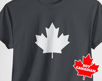 Puede incluir: Una camiseta gris oscuro con un diseño de hoja de arce blanca. La camiseta tiene el texto "Comprar canadiense" en rojo sobre una hoja de arce blanca.