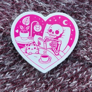May include: A pink heart-shaped sticker featuring a skeleton reading in bed with a cat, a plant, and a cup of coffee. The sticker has a white border and is set against a textured maroon background.