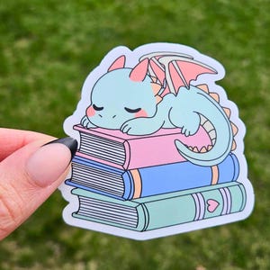 May include: A sticker featuring a cartoon dragon resting on a stack of books. The dragon is light blue with pink accents, and the books are pink, blue, and green. The sticker has a white border and is set against a green background.