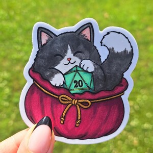 May include: A die-cut sticker featuring a black and white cat with a smiling expression, nestled inside a deep red drawstring pouch. The cat is holding a teal twenty-sided die with the number 20 clearly visible on its front face.