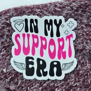 May include: A white sticker with the text "IN MY SUPPORT ERA" in black and pink. The sticker has a heart with a halo, wings, and cross symbols. The sticker is on a burgundy knitted surface.