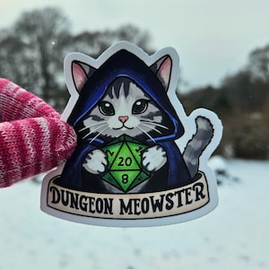 May include: A sticker featuring a cartoon cat wearing a blue hooded cloak and holding a green D20 die. The cat has gray and white fur and large green eyes. The sticker has the text "DUNGEON MEOWSTER" on a banner.