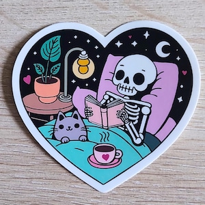 May include: A heart-shaped sticker with a skeleton reading a book in bed with a cat and a cup of coffee. The scene is set at night with stars and a crescent moon in the background.