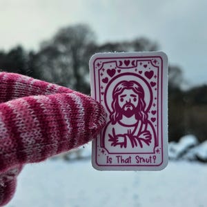 May include: A rectangular sticker featuring a magenta illustration of Jesus with a halo, surrounded by hearts and stars. The text "Is That Smut?" is at the bottom. The sticker is held by a pink and white striped glove against a snowy background.