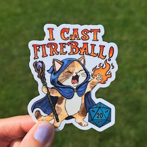 May include: A die-cut sticker featuring a calico cat wearing a blue hooded cloak, holding a wooden staff with a purple orb, and casting a flame from its paw. Below the cat is a blue twenty-sided die. The text I CAST FIREBALL! is above.
