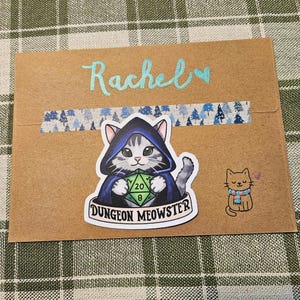 May include: A brown envelope with the name "Rachel" in light blue script. It features a "Dungeon Meowster" sticker of a cat in a hooded cloak holding a die, and a small cat sticker with a scarf.