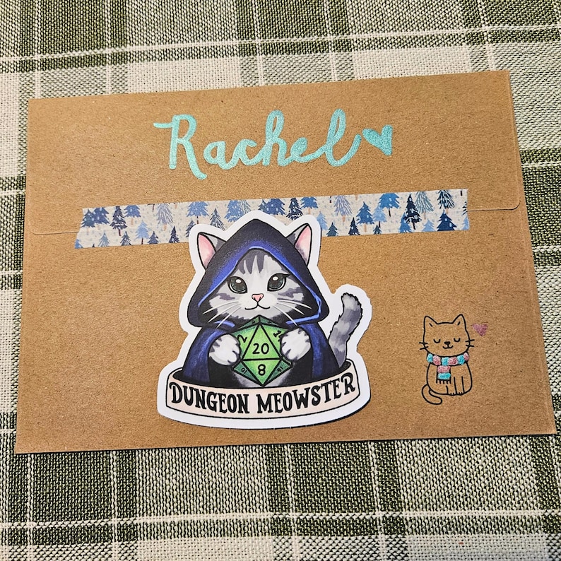 May include: A brown envelope with the name "Rachel" in light blue script. It features a "Dungeon Meowster" sticker of a cat in a hooded cloak holding a die, and a small cat sticker with a scarf.