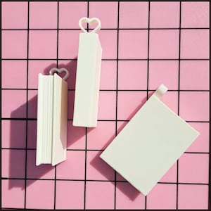 May include: Three off-white, solid-colored book-shaped objects made of a smooth material. Two of the items feature a small, hollow heart-shaped loop at the top, while the third item is a rectangular block with a flat top edge and no loop.