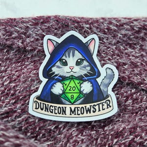 May include: A sticker featuring a gray tabby cat wearing a blue hooded cloak and holding a green and black twenty-sided die. The sticker has the text "DUNGEON MEOWSTER" on a banner.