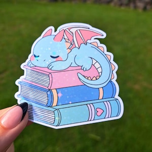 May include: A holographic sticker featuring a light blue dragon with pink wings sleeping on a stack of three books. The top book is pink, the middle is dark blue with stars, and the bottom is light blue with a pink heart detail.