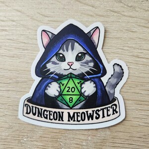 May include: A sticker featuring a gray and white cat wearing a blue hooded cloak and holding a green D20 die. The sticker has the text "Dungeon Meowster" on a banner below the cat.