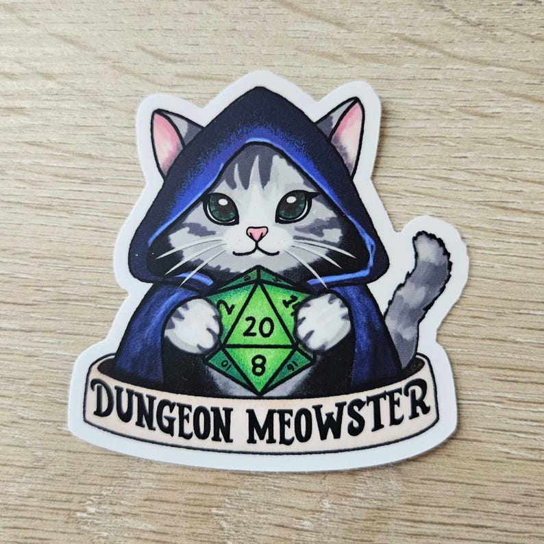 May include: A sticker featuring a gray and white cat wearing a blue hooded cloak and holding a green D20 die. The sticker has the text "Dungeon Meowster" on a banner below the cat.