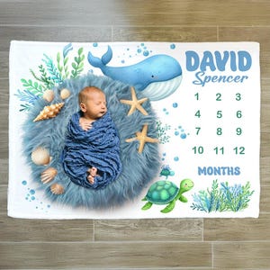 May include: A white baby blanket with a blue whale, starfish, seashells, and a green sea turtle. The blanket also has a blue and white fur rug with a baby sleeping on it. The blanket has the text "DAVID Spencer" and a monthly growth chart.