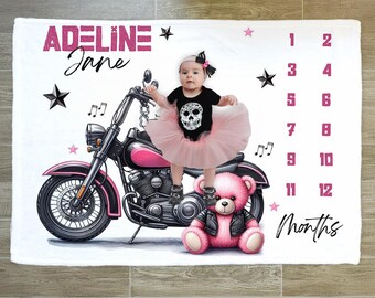 May include: A white baby milestone blanket featuring a pink and black motorcycle illustration. The blanket includes the name "Adeline Jane" in pink, along with star and musical note graphics. A baby in a tutu and a teddy bear are also featured.