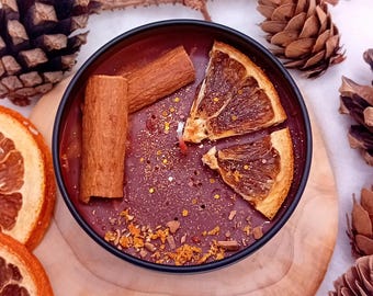 Può includere: Una candela profumata in una scatola nera, decorata con fette d'arancia essiccate e bastoncini di cannella. La candela è posizionata su un sottobicchiere di legno, circondata da pigne e un rametto. La candela ha una cera marrone rossiccio.