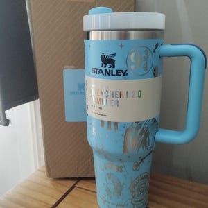 Puede incluir: Una taza de viaje de acero inoxidable azul con un asa negra. La taza tiene una tapa blanca y un diseño plateado con el texto "Stanley" y "9 3/4".
