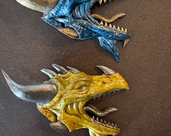 Puede incluir: Dos esculturas de cabeza de dragón, una azul y otra amarilla, con la boca abierta y dientes afilados. Ambas esculturas tienen escamas y cuernos detallados.