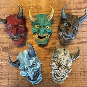 Könnte beinhalten: Fünf farbenfrohe japanische Dämonenmasken mit Hörnern. Die Masken sind aus bemaltem Gips gefertigt und haben unterschiedliche Gesichtsausdrücke. Die Masken sind in einer Reihe auf einer Holzoberfläche angeordnet.