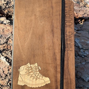 May include: A wooden hiking journal featuring a laser-etched design of hiking boots. The journal has the words "HIKING JOURNAL" engraved on the side and a black elastic band closure. The wood has a warm, natural tone.