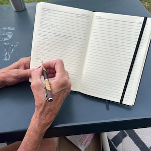 May include: A black and white notebook with a black elastic band closure. The notebook is open to a page with a list of categories for camping trip planning. A person is writing in the notebook with a brown and silver pen.