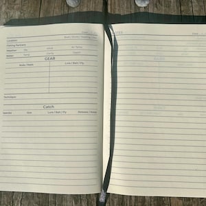 May include: A black and white fishing log book with sections for location, weather, gear, catch, and notes. The book has lined pages for writing.