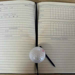 May include: A golf scorecard book with a white golf ball and a black tee on the right page. The left page has a section for recording golf course information, including location, tournament, event name, round, course, weather, tee time, partner, group, and a summary section with categories for eagles, birdies, par, bogey, double bogey, and putts per green. The right page has a grid for recording scores for front 9 and back 9 holes, with columns for par, fairway hit, GIR, total putts, and score. The bottom of the right page has a section for additional notes.